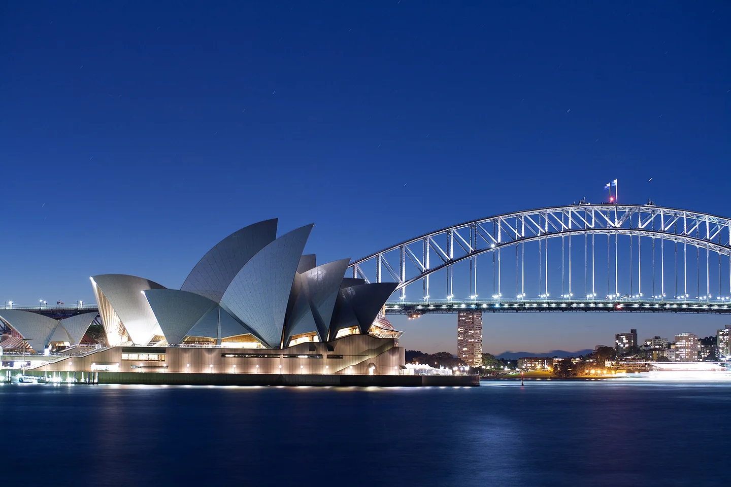The illuminated white sails of Sydney Opera House and the Harbour Bridge reflect in the water at blue hour.