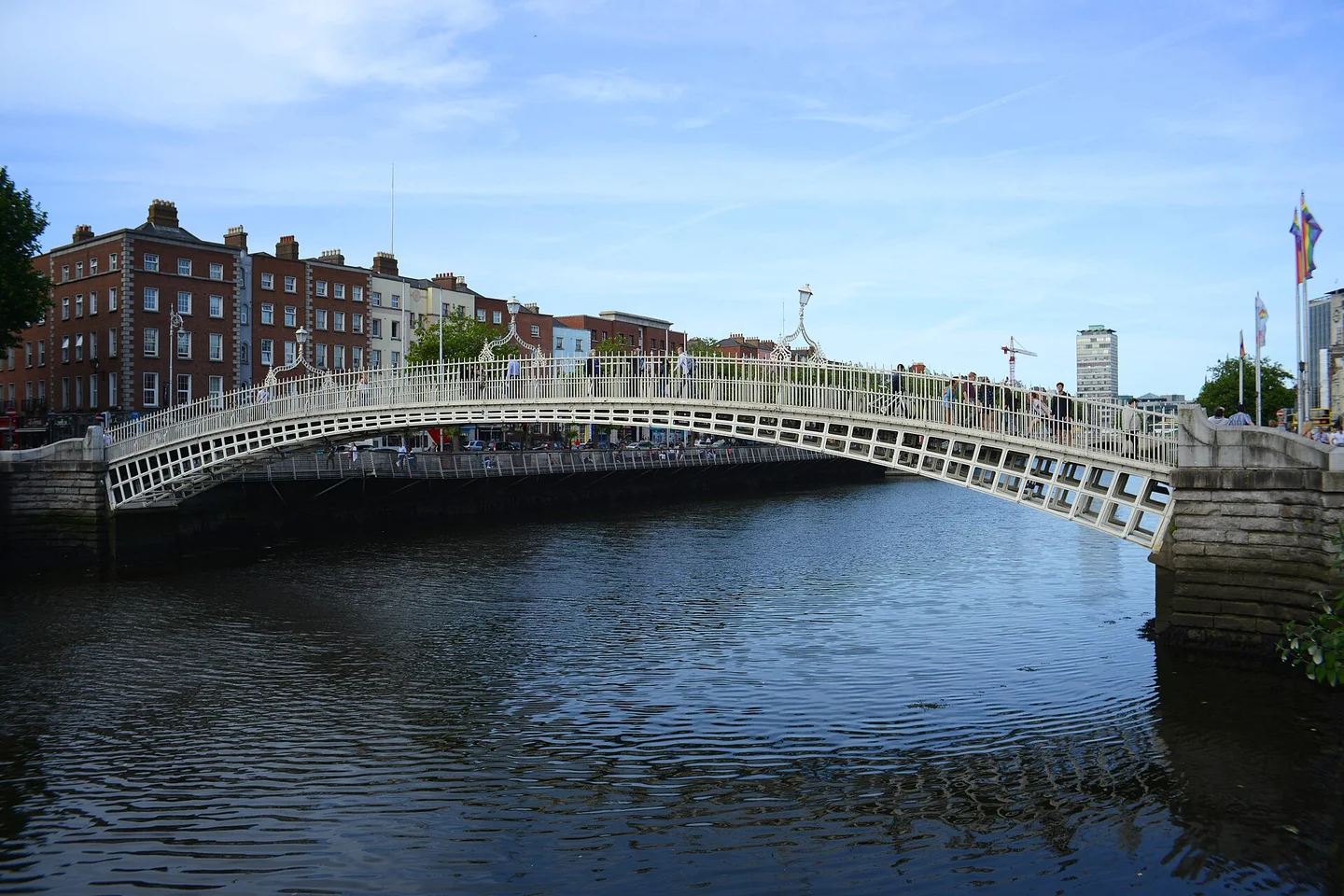 The white iron arches of Ha'penny Bridge cross the River Liffey beside Dublin's riverside buildings under a blue sky.