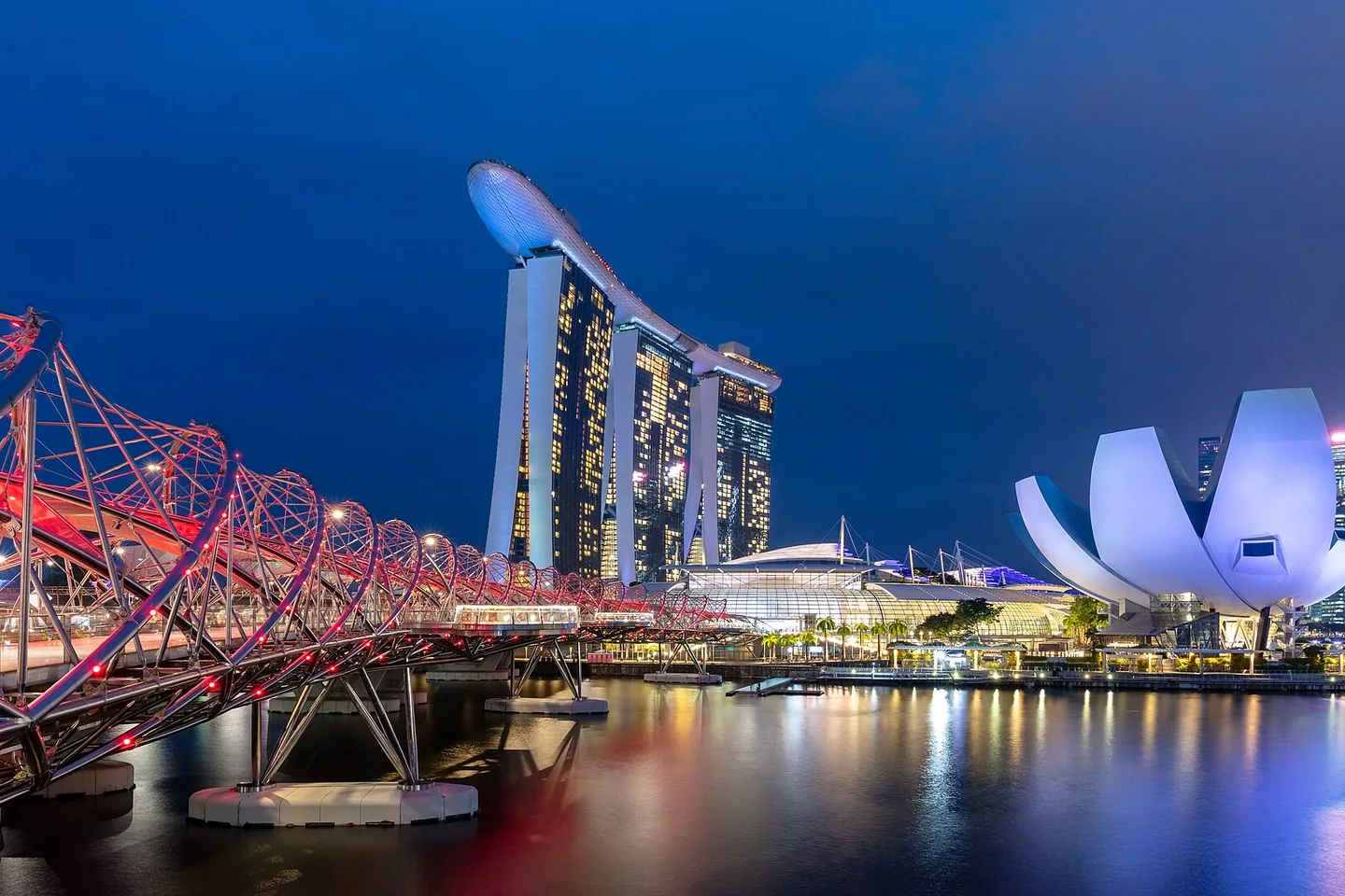 The illuminated Helix Bridge, ArtScience Museum and Marina Bay Sands against a blue evening sky.
