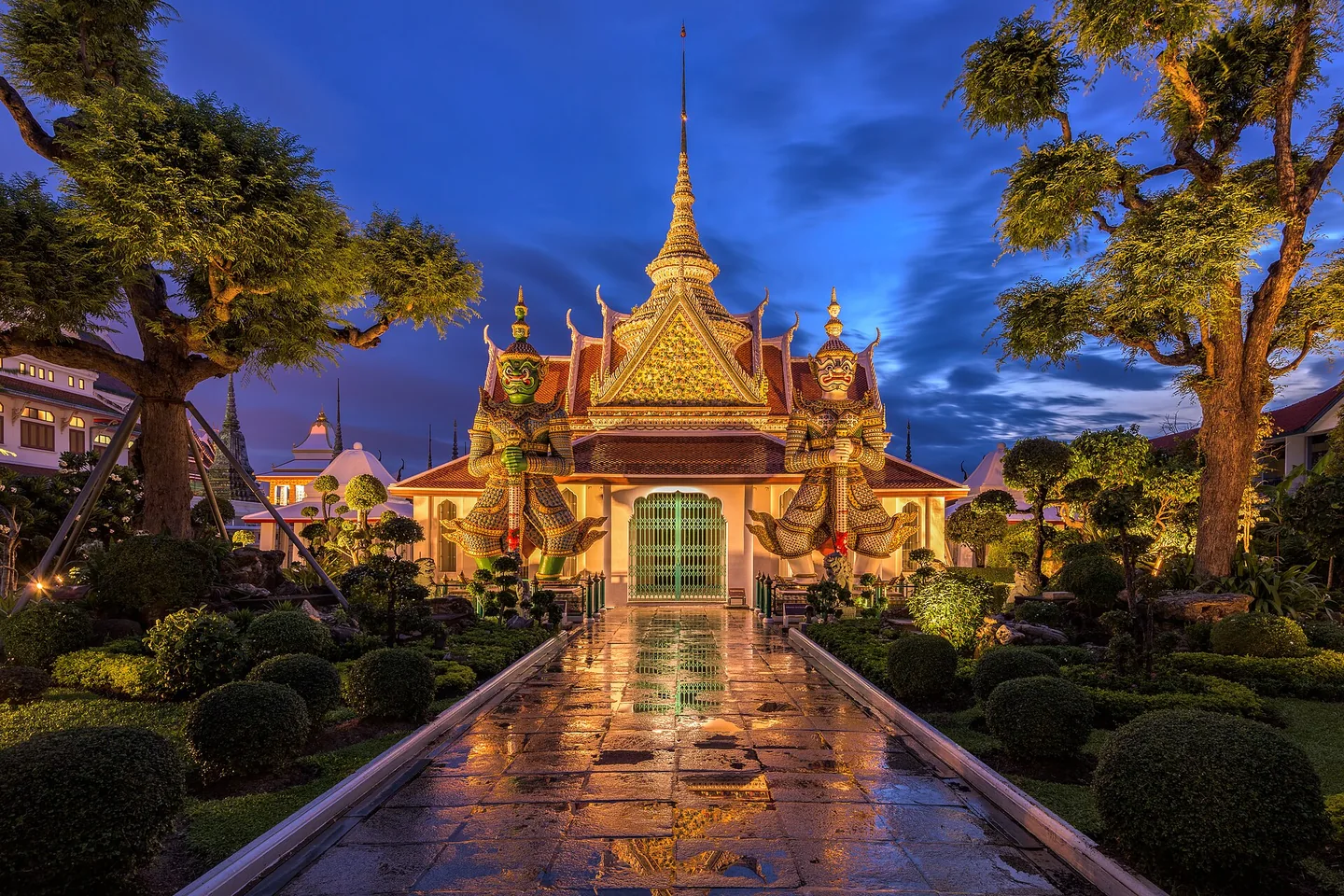 The ornate golden temple gateway and gardens at Wat Arun under a deep blue evening sky.