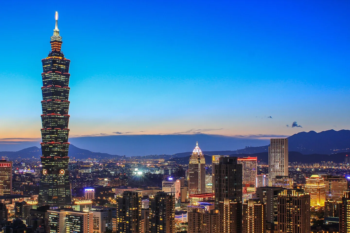 Taipei 101 rises over a glowing city skyline beneath a vivid blue evening sky.