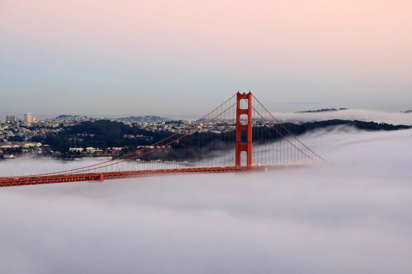 The red towers of the Golden Gate Bridge rise above low fog beneath a pink evening sky in San Francisco.