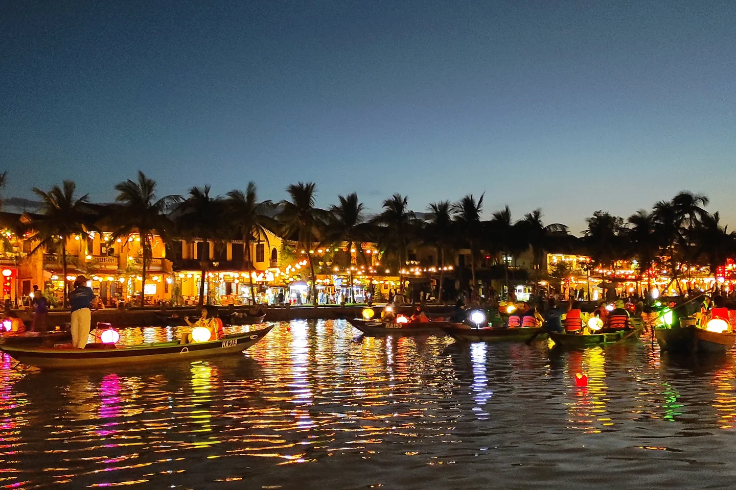 Lantern-lit riverside buildings and boats reflected in the Thu Bồn River at dusk in Hội An.